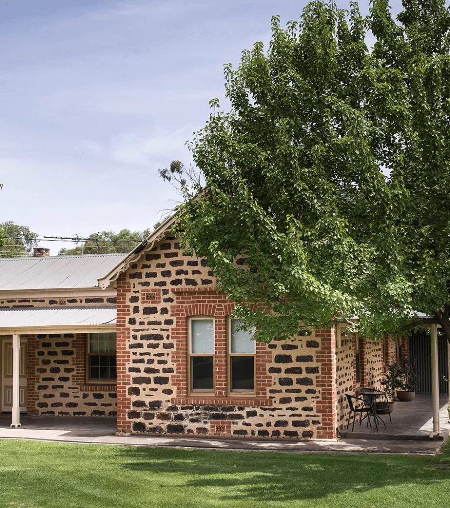 Exterior of a historic stone building with veranda, tree shade, and green lawn